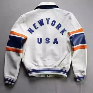Puede incluir: Chaqueta de cuero blanca con detalles azules y naranjas. La parte trasera muestra el texto "NEW YORK USA" en azul. Las mangas tienen rayas azules y naranjas. Estilo universitario.