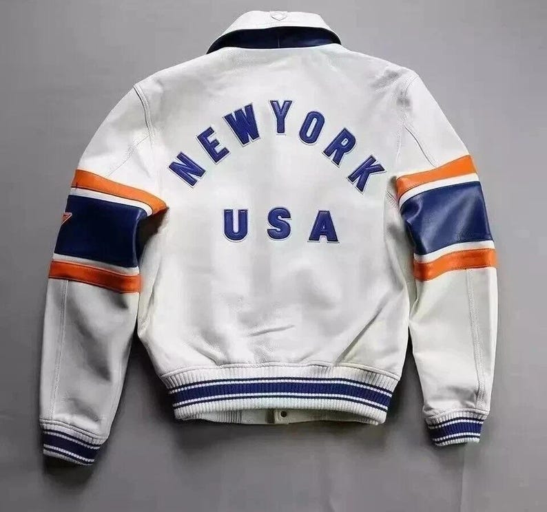 Puede incluir: Chaqueta de cuero blanca con detalles azules y naranjas. La parte trasera muestra el texto "NEW YORK USA" en azul. Las mangas tienen rayas azules y naranjas. Estilo universitario.