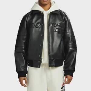 May include: A black leather varsity jacket featuring a pointed collar, snap button closure, and ribbed cuffs. The chest displays embossed Avirex branding. The sleeves include textured leather panels, and the jacket is worn over a cream hoodie.