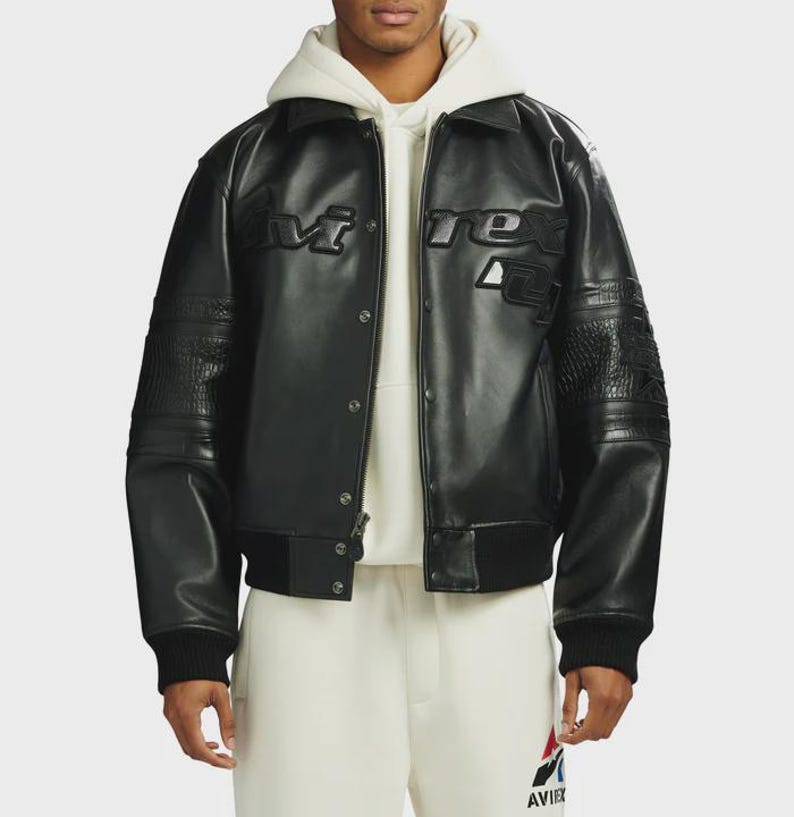 May include: A black leather varsity jacket featuring a pointed collar, snap button closure, and ribbed cuffs. The chest displays embossed Avirex branding. The sleeves include textured leather panels, and the jacket is worn over a cream hoodie.