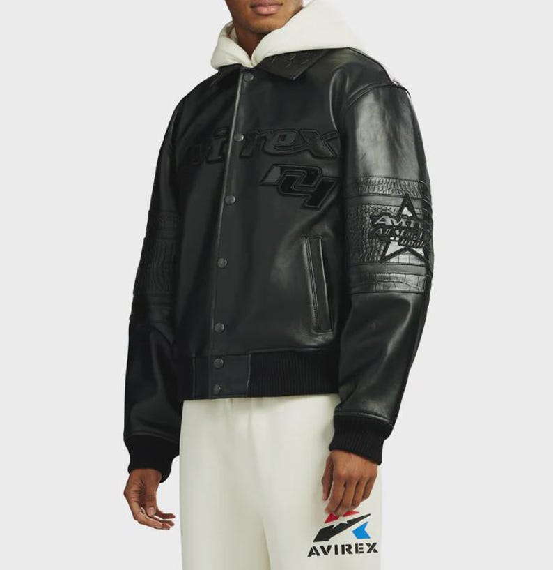 May include: A black leather varsity jacket featuring a snap-button front closure, a ribbed hem, and cuffs. The chest displays embossed Avirex branding, while the sleeves feature textured panels and a star graphic with the text All American.