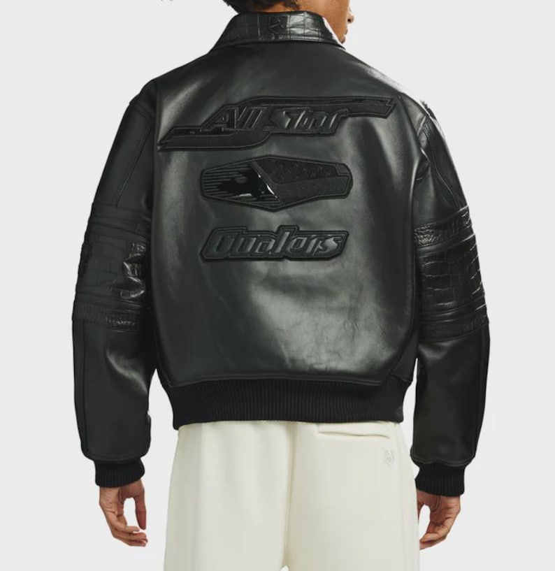 May include: A black leather bomber jacket featuring a ribbed collar, cuffs, and hem. The back displays embossed black patches with the text "ALL STAR" and "Qualers" in a stylized font, along with textured crocodile-patterned leather sleeves.