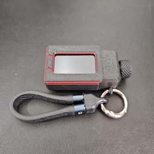 May include: A black and red digital device with a screen, attached to a black leather loop and silver keyring. The device has a textured surface and a red border around the screen. The keyring is attached to a black leather loop.