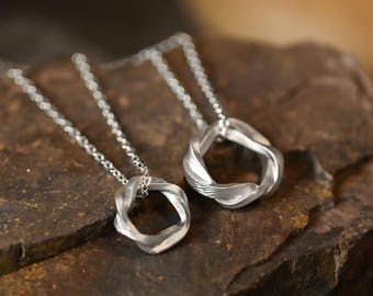 Puede incluir: Dos collares con colgante plateados. Cada collar presenta un diseño de círculo abierto y retorcido. Los colgantes cuelgan de delicadas cadenas plateadas. Los collares se exhiben sobre una superficie de piedra marrón rugosa.