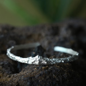 Puede incluir: Un brazalete plateado con un diseño de cordillera. El brazalete tiene una apariencia texturizada y rugosa, con un pico de montaña detallado en el centro. El fondo está borroso, con una superficie oscura y texturizada.