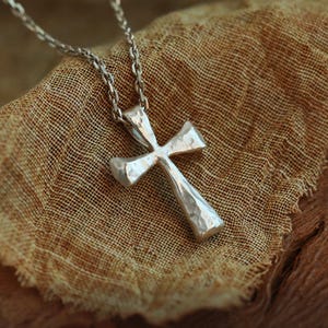 Peut inclure: Un collier pendentif argenté avec un motif de croix texturée. La croix est suspendue à une chaîne délicate. Le collier est présenté sur un tissu texturé de couleur beige.