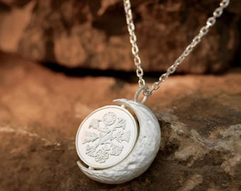 May include: A silver-toned pendant necklace featuring a crescent moon design. The moon holds a circular coin with a floral pattern. The necklace chain is delicate and the pendant is set against a blurred, textured background.