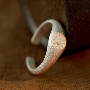 May include: A silver-colored ring with a textured surface and a sunburst design. The ring has an open design, allowing for adjustable sizing. The sunburst is etched into the metal, adding a decorative element to the jewelry.