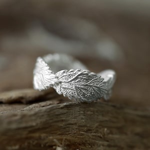 Puede incluir: Un anillo de plata que presenta una banda continua de hojas texturizadas y superpuestas. El metal tiene un acabado mate y cepillado que resalta las venas intrincadas y los bordes dentados de cada diseño de hoja que rodea todo el anillo.