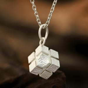 May include: A silver-colored pendant necklace featuring a cube-shaped charm. The charm is composed of multiple textured squares, suspended from a silver chain. The background is blurred, with a focus on the jewelry.