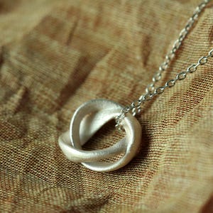 May include: A silver-toned pendant featuring two interlocking, textured circular rings suspended from a delicate matching metal chain. The pendant has a matte, slightly granular surface finish and a continuous, fluid design that creates a knot-like appearance.