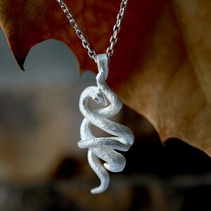 Op de afbeelding: Een zilverkleurige hangerketting met een ontwerp van een opgerolde slang. De slang is gedetailleerd met een getextureerd oppervlak en een vloeiende vorm. De hanger hangt aan een fijne zilveren ketting.