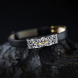 May include: A black leather bracelet featuring a rectangular silver-toned metal centerpiece with intricate geometric engravings and two small, inlaid gold-toned accents that resemble eyes.