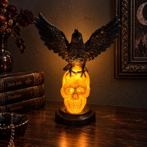 May include: A black raven figurine with outstretched wings perched atop a glowing, translucent yellow skull. The skull serves as a lamp base, emitting a warm light from within, and is mounted on a circular black stand.