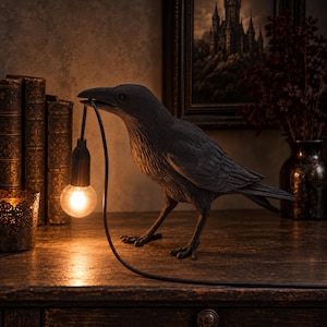 May include: A black raven figurine standing on a surface, holding a thin black cord in its beak that suspends a single glowing light bulb. The raven has detailed textured feathers and is positioned in profile, facing left with its head slightly lowered.