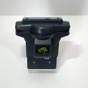 Puede incluir: Un dispositivo electrónico gris oscuro con una base rectangular y una parte superior curvada. El dispositivo tiene una pequeña pantalla y un logotipo verde y dorado. El dispositivo está sobre una superficie blanca.