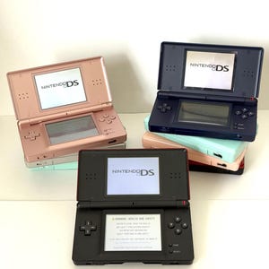 Puede incluir: Varias consolas portátiles Nintendo DS en tonos rosa, negro y azul marino. Cada consola está abierta, mostrando el logotipo de Nintendo DS en la pantalla. Las consolas están apiladas y dispuestas, mostrando su diseño.