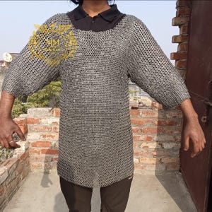 May include: A silver chainmail shirt with three-quarter sleeves. The shirt is made of small, interlocking metal rings. The person is wearing a black collared shirt underneath. The background includes a brick wall and a door.