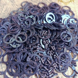 Puede incluir: Un montón de arandelas y remaches de metal de color bronce oscuro. Las arandelas son circulares con múltiples agujeros, y los remaches son pequeños sujetadores cilíndricos. Los artículos están esparcidos sobre una superficie marrón claro.