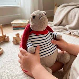 May include: A plush toy in the shape of a bulldog, wearing a striped shirt and a red scarf. The toy is held by a child's hands. The bulldog has a red cape and is surrounded by other toys.