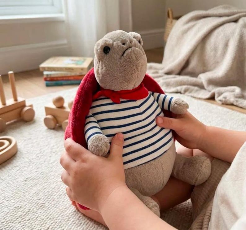 May include: A plush toy in the shape of a bulldog, wearing a striped shirt and a red scarf. The toy is held by a child's hands. The bulldog has a red cape and is surrounded by other toys.