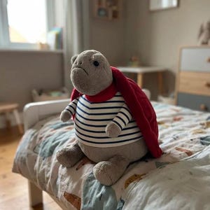 May include: A plush turtle toy with a gray body, wearing a striped shirt with navy blue and white stripes, a red cape, and a red scarf. The toy is sitting on a bed with a patterned comforter. The room has a window and a wooden dresser.