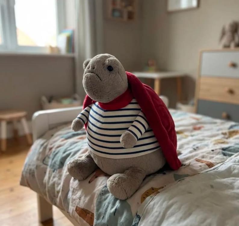 May include: A plush turtle toy with a gray body, wearing a striped shirt with navy blue and white stripes, a red cape, and a red scarf. The toy is sitting on a bed with a patterned comforter. The room has a window and a wooden dresser.
