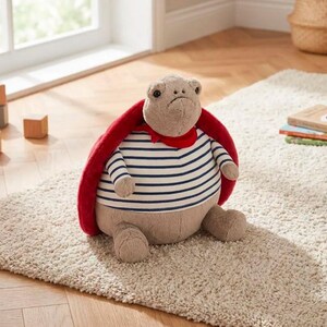 May include: A plush turtle toy with a red cape and a striped shirt. The turtle has a sad expression and is sitting on a beige rug. The toy is designed with soft, neutral colors.
