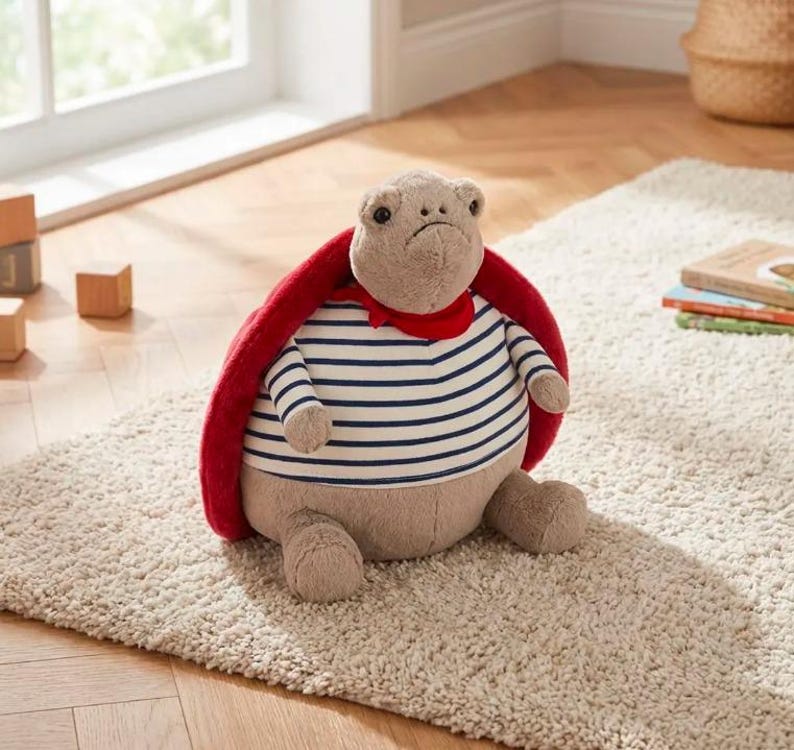 May include: A plush turtle toy with a red cape and a striped shirt. The turtle has a sad expression and is sitting on a beige rug. The toy is designed with soft, neutral colors.