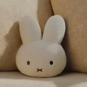 May include: A white plush bunny-shaped pillow with black dot eyes and a black 'x' for a mouth. The pillow has long, upright ears and is resting on a beige surface. The soft toy is designed to resemble a cartoon character.