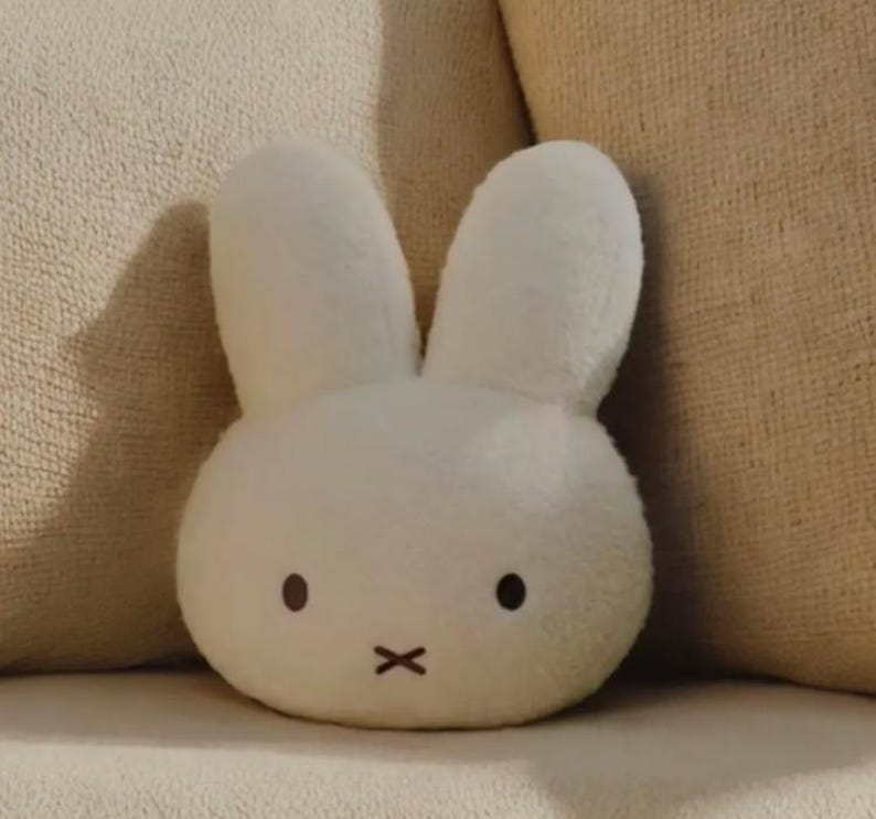 May include: A white plush bunny-shaped pillow with black dot eyes and a black 'x' for a mouth. The pillow has long, upright ears and is resting on a beige surface. The soft toy is designed to resemble a cartoon character.