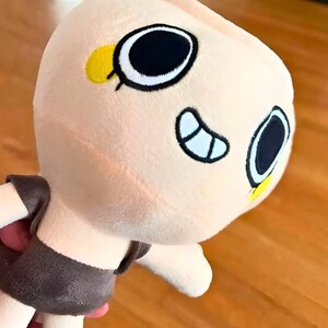 May include: A plush doll with a light peach-colored head and body. The doll has large, black and white eyes, a small yellow accent, and a brown outfit. The doll is soft and cuddly, perfect for children.