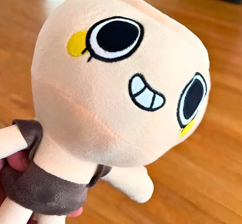 May include: A plush doll with a light peach-colored head and body. The doll has large, black and white eyes, a small yellow accent, and a brown outfit. The doll is soft and cuddly, perfect for children.