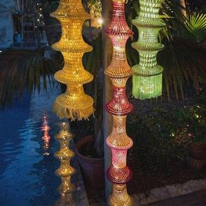 May include: Three cylindrical hanging lanterns featuring intricate crochet patterns in yellow, pink, and green. Each lantern has multiple tiered sections and a fringed bottom edge, with internal lights illuminating the woven structures.