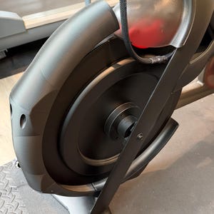 Pode incluir: Close-up do volante e da estrutura pretos de uma bicicleta de exercício. A bicicleta tem um design elegante e moderno com um acabamento escuro. A imagem destaca a roda, a estrutura e uma parte do assento da bicicleta, mostrando sua construção robusta e design.