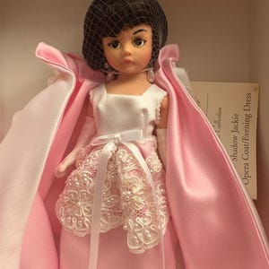 May include: A vintage doll wearing a white dress with a pink satin opera coat and a black net veil. The doll's dress features a white lace overlay with pearl accents and a white satin ribbon. The doll is part of the Shadow Jackie Collection.