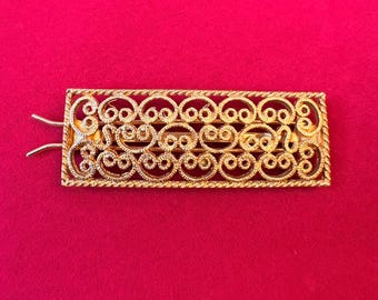 May include: A rectangular gold-tone hair barrette featuring an intricate openwork filigree design with repeating scroll patterns. The frame of the barrette has a twisted rope texture, and the metal surface has a polished, reflective finish.