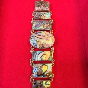 Op de afbeelding: Een schakelarmband met zes rechthoekige en één centraal ovaal abalone schelp-inlegstuk in zilverkleurig metaal. De iriserende schelpsegmenten vertonen wervelende patronen in groen, blauw, paars en goud. Aan de bovenkant bevindt zich een ronde sluiting.