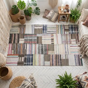 May include: A rectangular patchwork rug with a mix of neutral colors and vertical stripes. The rug features squares of various sizes and patterns, including blue, pink, and gray stripes. The rug is placed on a white wooden floor.