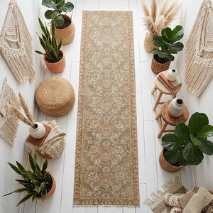 May include: A long, rectangular rug with a floral pattern in shades of green and peach. The rug is surrounded by potted plants, decorative macrame wall hangings, and small wooden stools. The overall aesthetic is bohemian and natural.