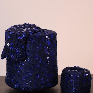 May include: A large cylindrical spool of dark navy blue yarn adorned with reflective blue sequins, accompanied by a smaller, separate ball of the same sequined navy blue yarn. The yarn features a consistent texture with evenly distributed sequins.