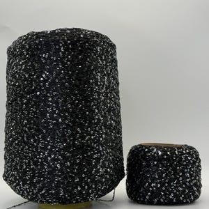 May include: A large cylindrical cone and a smaller spool of black yarn featuring a dense distribution of small, reflective silver sequins throughout the thread. The yarn has a textured, net-like appearance with metallic accents visible across both items.