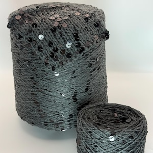 May include: A large cylindrical spool of dark gray yarn and a smaller matching ball of yarn. Both items feature a knitted texture integrated with numerous small, flat, reflective black sequins scattered throughout the dark gray thread.