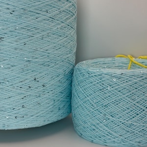 May include: Two cylindrical spools of light blue yarn featuring small, reflective silver sequins embedded throughout the thread. The larger spool stands to the left, while a smaller spool sits to the right with a bright yellow string on top.
