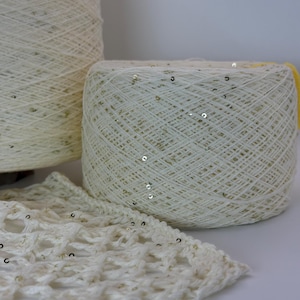 Op de afbeelding: Twee grote klossen off-white garen met kleine, reflecterende gouden lovertjes verwerkt in de draad. Een staal van opengewerkt haakwerk gemaakt van hetzelfde garen met lovertjes ligt op de voorgrond en toont de textuur van het materiaal.