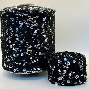 May include: A large spool and a smaller ball of black yarn featuring embedded iridescent silver sequins. The yarn is wound tightly, showcasing a consistent distribution of reflective, circular sequins throughout the dark, textured black thread.
