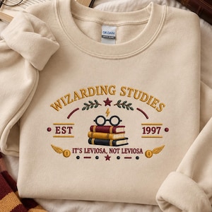 May include: A beige crewneck sweatshirt featuring an embroidered design with the text WIZARDING STUDIES, EST 1997, and IT'S LEVIOSA, NOT LEVIOSA. The graphic includes a stack of three books, round glasses, a lightning bolt, and two golden wings.