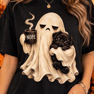 Op de afbeelding: Een zwart t-shirt met een afbeelding van een beige geest die een zwarte mok vasthoudt met de tekst NOPE in het wit gedrukt. De geest houdt ook een kleine zwarte mopshond vast. Het ontwerp is gecentreerd op de voorkant van het shirt en toont een Halloween-illustratie met een neutraal kleurenpalet.