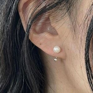May include: A close-up of a pearl earring on an ear. The earring features a small, round, white pearl stud and a dangling silver ball. The skin tone is fair, and the hair is dark.