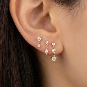 May include: A close-up of three sets of gold earrings with clear, sparkling gemstones. The earrings are in a variety of designs, including studs, dangling teardrops, and heart shapes. The earrings are worn on an ear.
